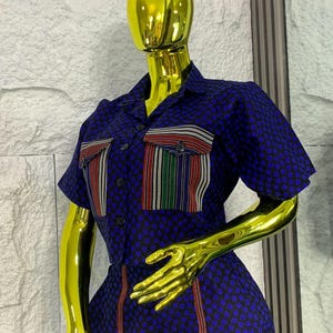 May include: A blue jumpsuit with a small floral pattern. The short-sleeved jumpsuit features two chest pockets with striped accents in red, white, and green. Vertical red stripes run down the sides of the legs. The outfit is displayed on a gold mannequin.