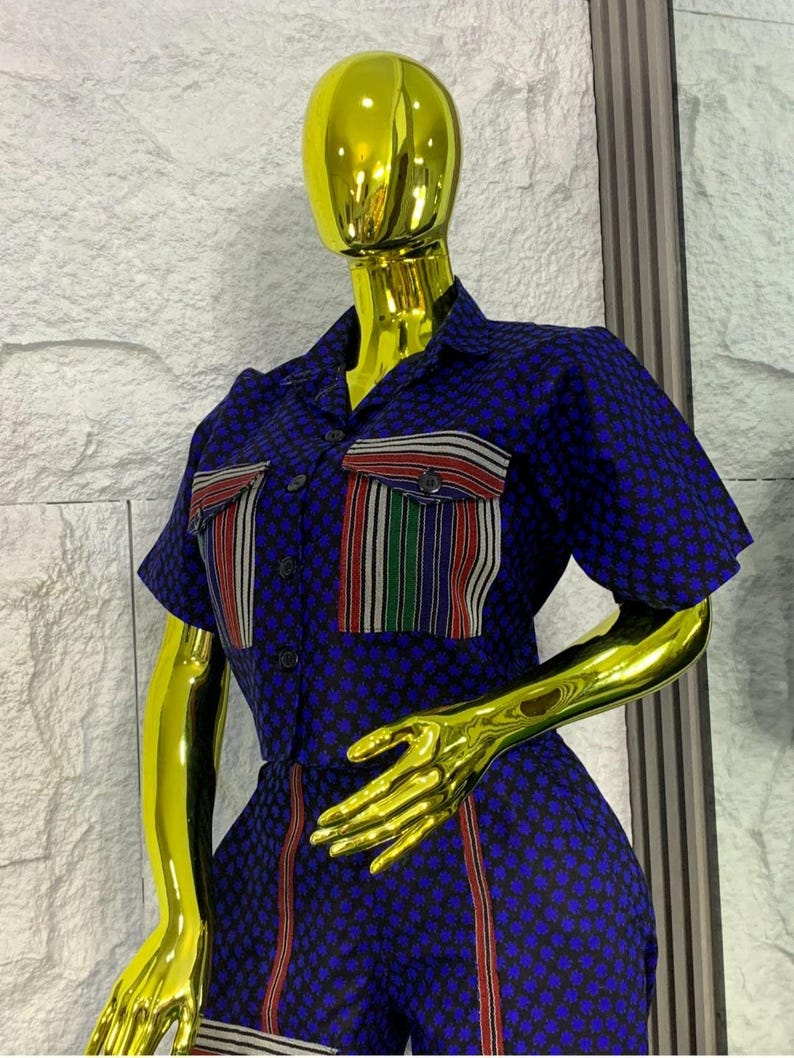 May include: A blue jumpsuit with a small floral pattern. The short-sleeved jumpsuit features two chest pockets with striped accents in red, white, and green. Vertical red stripes run down the sides of the legs. The outfit is displayed on a gold mannequin.