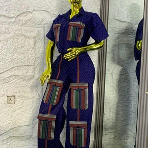 May include: A navy blue two-piece outfit displayed on a mannequin. The set comprises a short-sleeved shirt with a collar and wide-leg trousers. Both garments have a small, repeating pattern. The shirt and trousers have pockets with colourful striped details.
