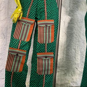 May include: Green patterned pants with four orange and striped pockets. The pants have a repeating clover-like pattern and brown vertical stripes. The pockets have a button closure. The outfit is displayed on a mannequin.