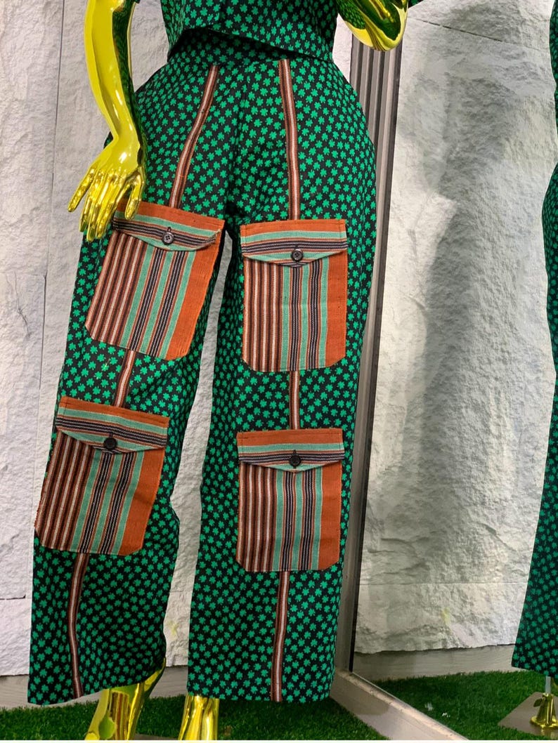 May include: Green patterned pants with four orange and striped pockets. The pants have a repeating clover-like pattern and brown vertical stripes. The pockets have a button closure. The outfit is displayed on a mannequin.