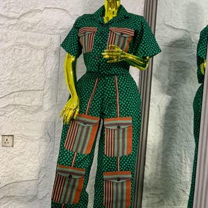 May include: Green jumpsuit with a polka dot pattern and short sleeves. The jumpsuit features four large pockets with a striped design in shades of orange, brown, and green. The outfit is displayed on a gold mannequin.