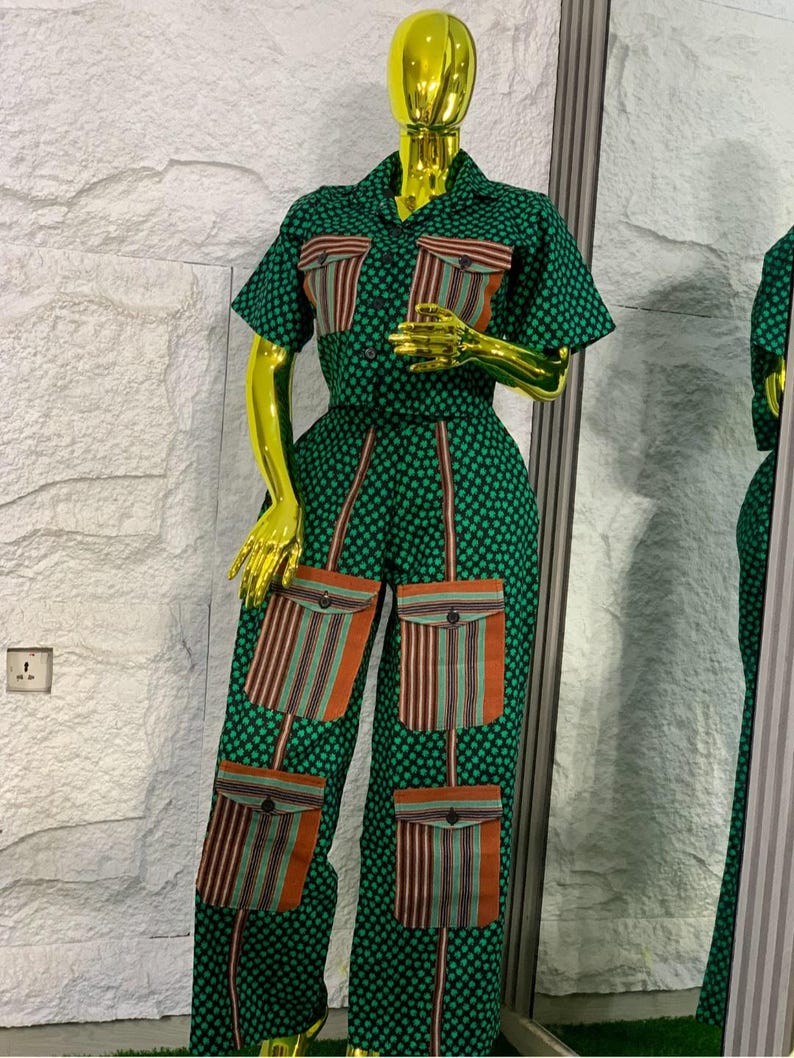May include: Green jumpsuit with a polka dot pattern and short sleeves. The jumpsuit features four large pockets with a striped design in shades of orange, brown, and green. The outfit is displayed on a gold mannequin.
