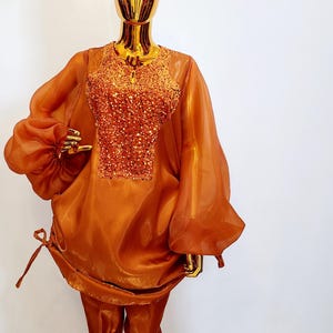 Peut inclure: Une tenue deux pièces de couleur orange brûlée. Le haut a des manches longues avec un détail plissé au poignet et un panneau pailleté au centre. Le pantalon est à jambe droite et a un léger brillant.
