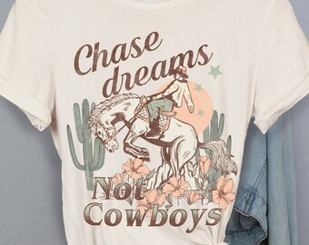 May include: Cream-colored t-shirt with a graphic of a cowboy riding a horse, surrounded by cacti and flowers. The text "Chase dreams Not Cowboys" is printed in brown. A denim jacket hangs in the background.