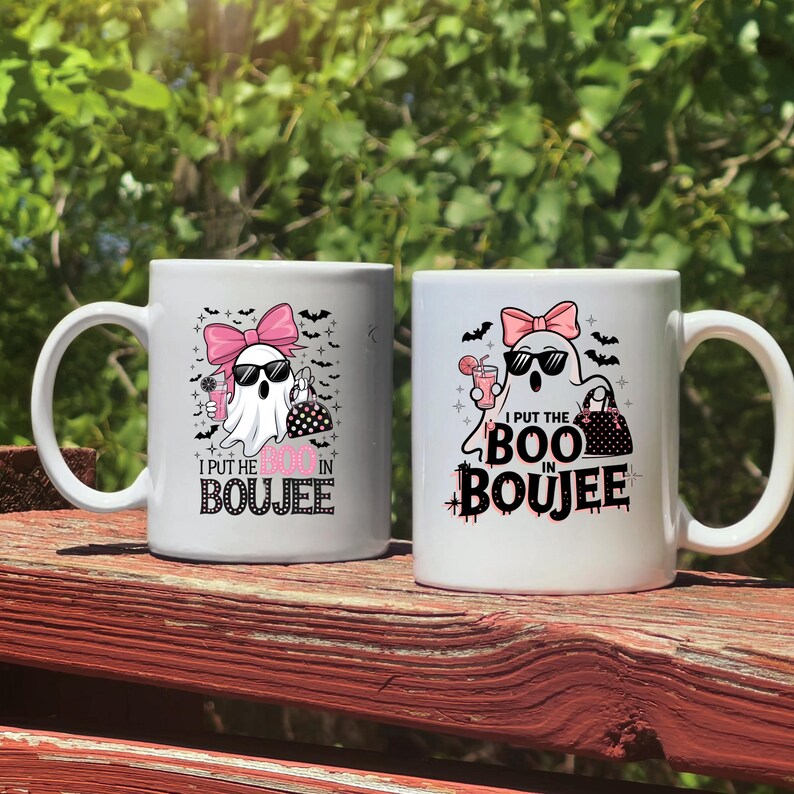 May include: Two white ceramic mugs with a black and white design of a ghost wearing sunglasses and a pink bow. The ghost is holding a pink drink and a black and white polka dot purse. The text on the mug reads "I put the boo in boujee".