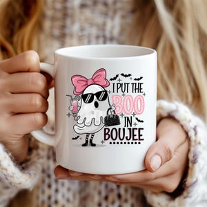 May include: White ceramic coffee mug with a pink ghost wearing sunglasses and a pink bow. The ghost is holding a purse and a pink drink. The text on the mug reads "I put the boo in boujee".