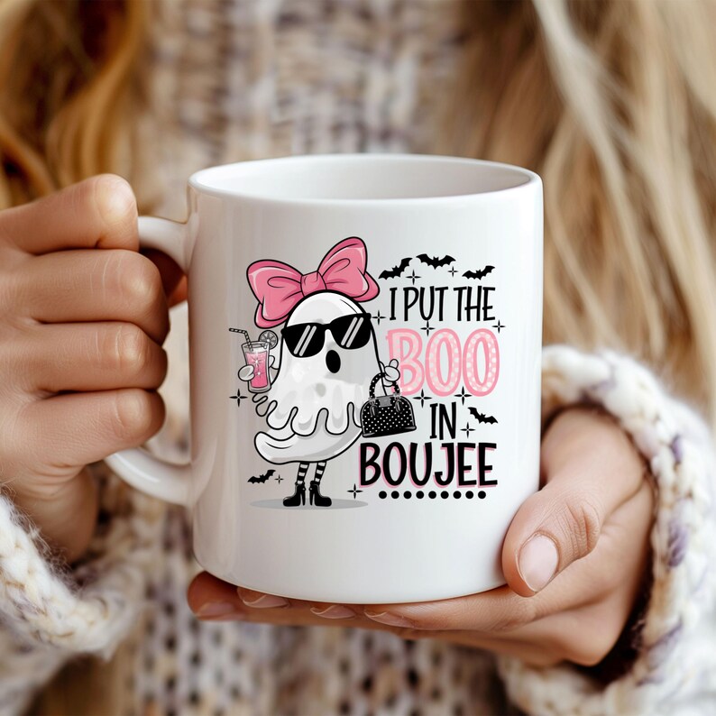May include: White ceramic coffee mug with a pink ghost wearing sunglasses and a pink bow. The ghost is holding a purse and a pink drink. The text on the mug reads "I put the boo in boujee".