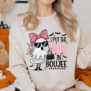 May include: A beige sweatshirt with a graphic of a ghost wearing sunglasses, a pink bow, and holding a drink and a purse. The text "I put the BOO in BOUJEE" is printed on the sweatshirt.