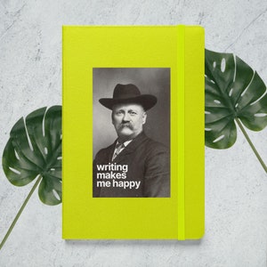Puede incluir: Un cuaderno amarillo brillante con un retrato de un hombre con sombrero y el texto "writing makes me happy". El cuaderno tiene un cierre de banda elástica amarilla a juego. Una hoja verde está en el fondo.