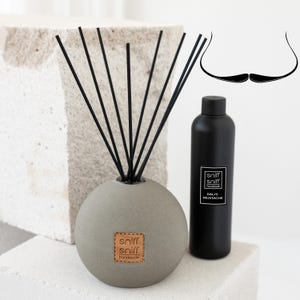 Könnte beinhalten: Eine graue Diffuserflasche mit schwarzen Schilfrohren und einem braunen Leder-Etikett mit der Aufschrift "sniff sniff handmade". Die Flasche steht auf einer weißen Oberfläche. Das Diffusionsöl befindet sich in einer schwarzen Flasche mit einem weißen Etikett mit der Aufschrift "sniff sniff handmade DALI'S MUSTACHE".