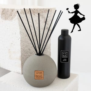 Könnte beinhalten: Ein runder, grauer Diffusor mit schwarzen Schilfrohren und einem braunen Leder-Etikett mit der Aufschrift "sniff sniff handmade". Der Diffusor steht neben einer schwarzen Flasche mit einem weißen Etikett, auf dem "sniff sniff handmade - AND JULIET" steht.