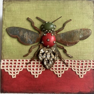 May include: A rusty metal bee with green and red jeweled accents, set against a green fabric background with a white lace trim and a red fabric base.