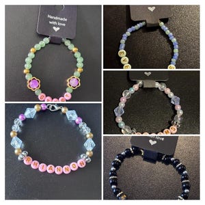 May include: A selection of handmade beaded bracelets. One bracelet has green beads, gold accents, and a flower design. Other bracelets include letter beads spelling out names. Each bracelet is attached to a tag that says "Handmade with love".