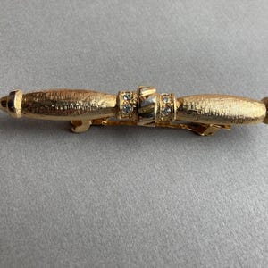 May include: Gold-toned barrette with a textured finish. The barrette features a central section adorned with small, clear rhinestones. The design includes rounded end pieces, creating a symmetrical and elegant accessory.