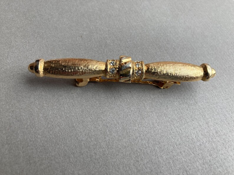 May include: Gold-toned barrette with a textured finish. The barrette features a central section adorned with small, clear rhinestones. The design includes rounded end pieces, creating a symmetrical and elegant accessory.