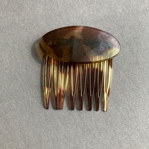 May include: A vintage hair comb with a tortoiseshell design. The comb features a curved, oval-shaped top and a series of thin, evenly spaced teeth. The colors range from light amber to dark brown.