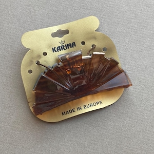 May include: A brown, fan-shaped hair claw clip on a gold-colored cardboard backing. The clip is made of translucent plastic and has a tortoiseshell pattern. The cardboard has the brand name "KARINA" and the text "MADE IN EUROPE".
