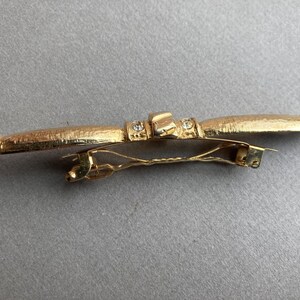 May include: A gold-toned barrette with a textured surface and a decorative design. The barrette features a central rectangular element with small, clear embellishments. The clasp is a simple, gold-colored metal.