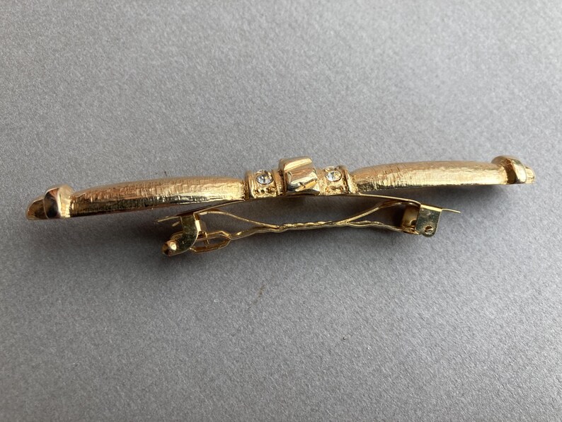 May include: A gold-toned barrette with a textured surface and a decorative design. The barrette features a central rectangular element with small, clear embellishments. The clasp is a simple, gold-colored metal.