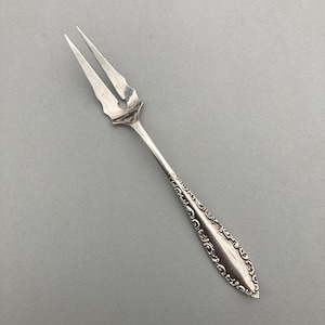 May include: A silver two-tined fork with a decorative handle. The fork's tines are sharp and pointed, and the handle features an ornate, engraved design. The fork is likely for serving or eating small portions.