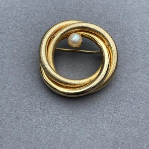 May include: Gold-toned brooch featuring a circular design with interwoven loops. A single, round pearl is centered within the loops. The brooch has a textured surface and a pin closure on the back. The jewelry piece is set against a gray background.
