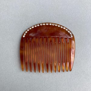 May include: A brown hair comb with a curved top, adorned with a row of small, clear rhinestones. The comb has a series of evenly spaced teeth. The comb is made of a translucent material.