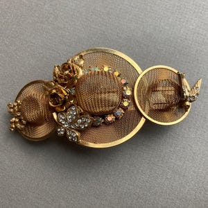 May include: Gold-toned metal hat-shaped brooch with mesh detailing. Features three hats, embellished with rhinestones, flowers, and a butterfly. A vintage-inspired accessory.