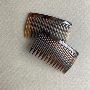 May include: Two tortoiseshell-patterned hair combs. The combs have a curved design with closely spaced teeth. The color transitions from a dark brown to a lighter amber hue at the tips of the teeth. The combs are likely made of plastic.