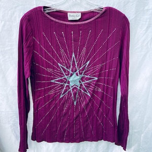 May include: A long-sleeved, plum-colored shirt with a starburst design. The shirt features a silver glitter star in the center, with lines of small silver dots radiating outwards. The text "Pretty Girl" is visible near the neckline.