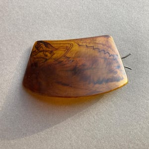 May include: A tortoiseshell-style hair clip with a curved rectangular shape. The clip features a dark brown and amber color scheme with a decorative design on the top surface and two metal prongs.