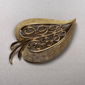May include: A gold-toned leaf-shaped brooch with a textured surface. The brooch features an openwork design with swirling, scroll-like details. The overall design is reminiscent of a stylized leaf.