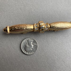 May include: Gold-toned bar pin with a textured finish. The pin features a central section adorned with clear rhinestones and two rounded ends. A US quarter is next to the pin for size comparison. Jewelry.