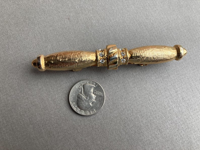 May include: Gold-toned bar pin with a textured finish. The pin features a central section adorned with clear rhinestones and two rounded ends. A US quarter is next to the pin for size comparison. Jewelry.