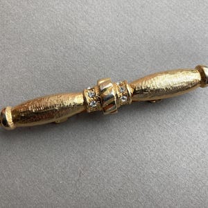 May include: A gold-toned bar pin with a textured surface. The pin features a central band adorned with small, clear crystals and rounded ends. The pin is approximately 3 inches long.