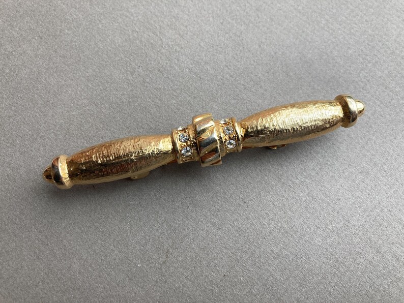 May include: A gold-toned bar pin with a textured surface. The pin features a central band adorned with small, clear crystals and rounded ends. The pin is approximately 3 inches long.