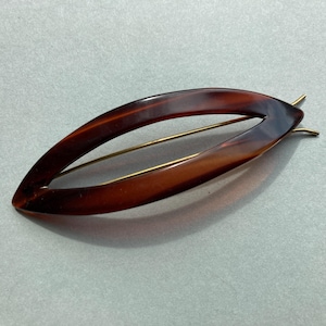 May include: A vintage hair clip in a leaf shape, made of tortoiseshell-patterned plastic. The clip features a gold-colored metal pin for securing it in the hair. The clip is approximately 4 inches long.