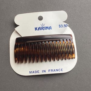 May include: A brown tortoiseshell hair comb on a white card with the brand name "Karina" and "Made in France" printed on it. The comb has fine teeth and a curved shape, suitable for styling hair.