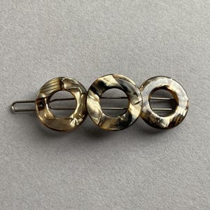 May include: A hair barrette with three circular, open-center designs. The barrette features a tortoiseshell pattern in shades of brown, tan, and black. The barrette is made of a shiny material and is attached to a silver-colored metal clip.