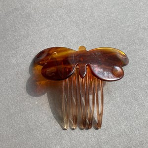 May include: A tortoiseshell-colored hair comb with a butterfly design. The comb features a butterfly-shaped top with a series of teeth extending downwards. The comb is made of a translucent material, and the butterfly design has a marbled appearance.