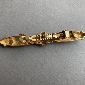 May include: A gold-toned metal barrette with an ornate design. The barrette features a rectangular shape with decorative embellishments on each end and a central clasp mechanism. The overall design is symmetrical and elegant.