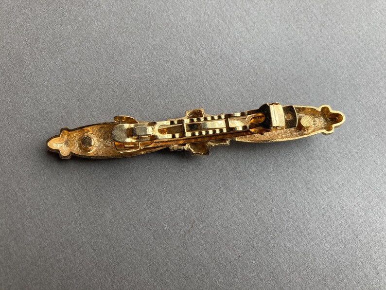 May include: A gold-toned metal barrette with an ornate design. The barrette features a rectangular shape with decorative embellishments on each end and a central clasp mechanism. The overall design is symmetrical and elegant.
