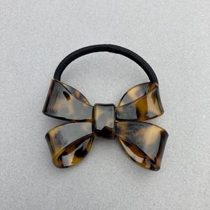 May include: A brown and black tortoiseshell hair tie with a large bow. The bow is made of plastic and has a glossy finish.