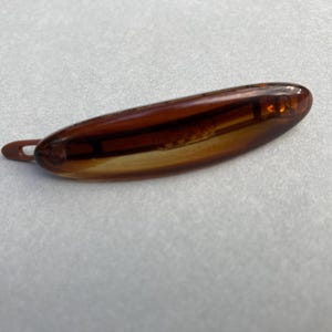 May include: A brown, oval-shaped hair clip with a small loop at one end. The clip is made of a translucent material, allowing light to pass through. The clip is a warm, amber color.