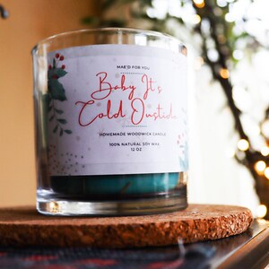 May include: A clear glass jar candle with a teal blue wax and a white label that reads "Baby It's Cold Outside" with a red and green holly design. The candle is on a cork coaster.