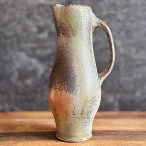 May include: A handmade ceramic pitcher with a handle. The pitcher has a light brown and green glaze with darker brown accents. The pitcher is on a wooden surface with a gray background.