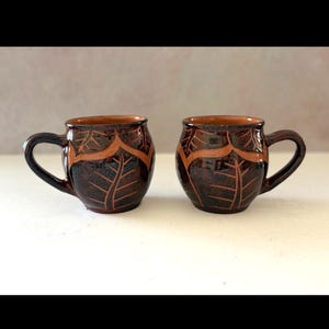 May include: Two rounded ceramic mugs featuring a dark brown, mottled glaze with carved leaf patterns. The design includes a lighter terracotta-colored band near the rim and base, with deep, etched lines forming leaf veins across the surface.