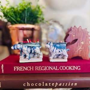 May include: Two ceramic match holders, each shaped like a dog, with blue and white detailing. The holders sit atop a red book titled "FRENCH REGIONAL COOKING". Red-tipped matches are visible inside the holders.