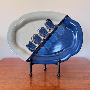 May include: An oval ceramic platter with a two-tone design. One half is light gray with blue floral accents, the other is a glossy dark blue. The platter is displayed on a black metal stand, showcasing its decorative appeal. A unique piece for serving or display.