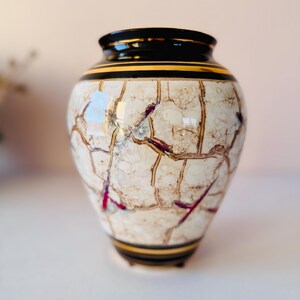 May include: A decorative ceramic vase with a cracked, off-white surface, accented with brown and red veining. The vase has a black rim with gold trim and a rounded shape, perfect for home decor.
